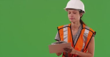 Kadın inşaat işçisi yeşil ekranda panoya notlar yazıyor. İnşaatçı kadın yeşil ekran duvarın önünde çelik yelek ve kask takıp panoya bakıyor. 4k