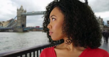Kasvetli görünümlü siyah kadın Londra, İngiltere 'de kameradan başka tarafa bakıyor. Düşünceli bir Afro-Amerikalı kadın, arka planda Tower Bridge ile ekranın dışına bakıyor. 4k