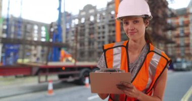 Kasketli ve güvenlik yeleği giyen kadın inşaat işçisi kameraya gülümseyen büyük bir iş yerinin yanında panoda duruyor. 4k