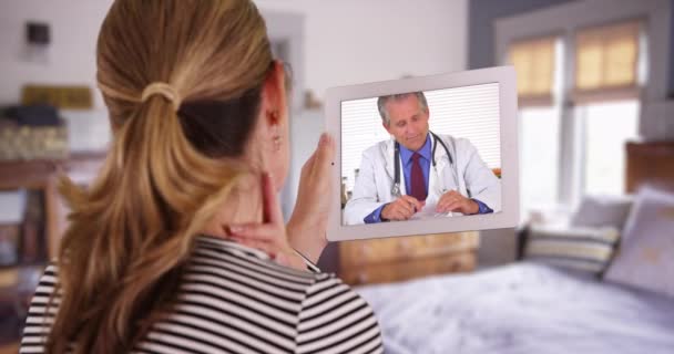 白人女性のビデオでは ベッドルーム内のタブレットで白人男性医師を呼んでいます 家庭からのビデオチャットを通じて医療専門家と話す後ろから示された女性または患者 — ストック動画