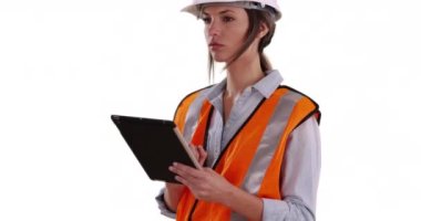 Turuncu yelek giyen ve beyaz arka planda tablet üzerinde çalışan bir kadın. Odaklanmış kadın inşaat işçisi beyaz fotokopi alanındaki ped cihazı üzerinde çalışıyor. 4k