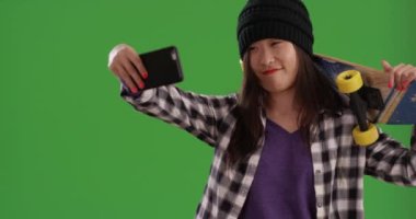 Genç Asyalı hippi yeşil ekranda akıllı telefonla selfie çekerken kaykayı omuzunda taşıyor. Rahat Çinli kaykaycı kız yeşil ekranda selfie çekmek için akıllı telefon kullanıyor. 4k