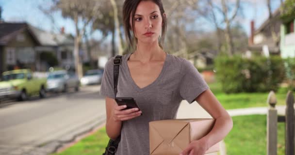 在住宅人行道上持邮包时使用手机的妇女 千禧年女性使用智能手机应用追踪郊区社区外的软件包 — 图库视频影像