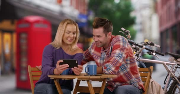 情侣们在伦敦街上的智能手机上用红色电话亭观看有趣的视频 快乐的男人和女人在户外喝咖啡放松 — 图库视频影像