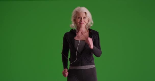 年配の女性は自信を持って迅速にグリーンスクリーン上のカメラに向かって歩いてください スポーツウェアやイヤーバッドを目的として走る高齢者の女性が鍵をかけられ または構成される — ストック動画