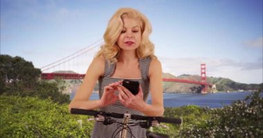 Neşeli Kafkasyalı kadın San Francisco 'da bisiklet sürerken cep telefonunu yol tarifi olarak kullanıyor. Mutlu turist, arka planda ünlü Golden Gate köprüsünü bulmak için akıllı telefon kullanıyor. 4k