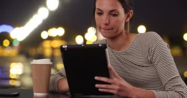 Milenyum kadını, şehir ortamında tabletle kafeterya masasında oturuyor. Yirmili yaşlarda bir kadın, kahve ve ped kullanıyor. Geceleri şehirde masada oturuyor. 4k