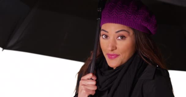 接近迷人的拉丁女性 戴着围巾和贝尼帽 在工作室里用雨伞遮挡雨 一个女人站在白色背景的黑色伞下的画像 — 图库视频影像