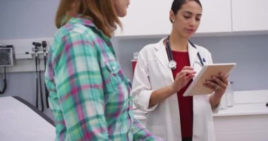 Hastanın sağlık durumunu not etmek için tablet kullanan genç ve güzel Latin doktorların portresi. Klinikteki genç tıp doktoruna danışmanlık yapan yaşlı kadın hastayı kapatın.