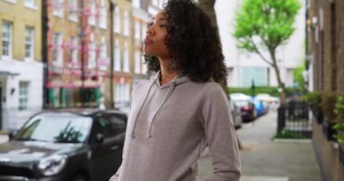Gri kapüşonlu siyahi kadın şehir kaldırımında gezinmeyi beklerken ileri geri sallanıyor. Güzel Afrikalı Amerikalı kız, şehirde taksi ya da araç paylaşımı bekliyor. 4k
