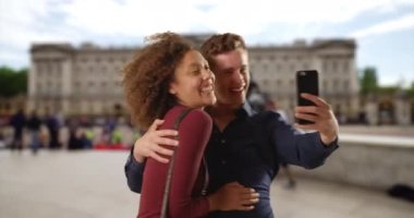 Neşeli çift Buckingham Sarayı yakınlarında akıllı telefonla fotoğraf çekiyor. Beyaz erkek ve kadın balayında Londra 'da selfie çekiyorlar. 4k
