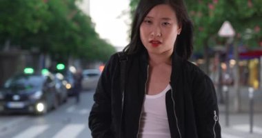 Şehir caddesinde rüzgarda uçuşan saçları olan siyah bombardıman ceketli Çinli kadın. Asyalı milenyum turisti Paris 'te cepleriyle kaldırımda bekliyor. 4k