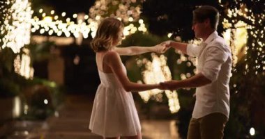 Dışarıda dans eden çekici genç bir çift akşamları ışıklara sarılıyor. Geceleri açık havada romantik erkek arkadaşıyla dans eden beyaz elbiseli, gülümseyen bir kadın. 4k