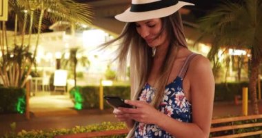 Çiçekli Romper ve fötr şapkalı mutlu bir kadın tropik bir otelde telefonla mesajlaşıyor. 20 'li yaşlarda bir kız tropikal ortamda tatildeyken cep telefonundan mesaj atıyor. 4k