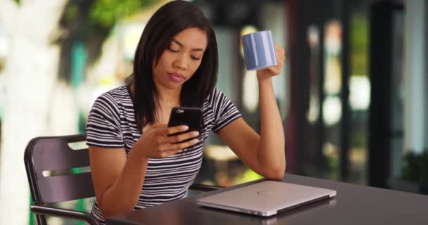 迷人的黑人女性坐在咖啡店外面 一边打滚一边打电话 笑着看社交媒体上的图片 非裔美国妇女拿着咖啡杯在手机上浏览 — 图库视频影像