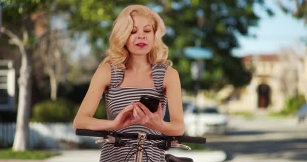 スマートフォンで郊外の通りのテキストメッセージに自転車で立って縞模様のサンドレスで魅力的な白人女性 日当たりの良い近所の携帯電話で自転車のメッセージングで休んでいる女性を笑顔 — ストック動画