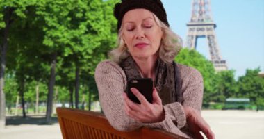 Emekli emekli Paris 'te tatilde. Park bankında oturmuş Eiffel Kulesi' nin akıllı telefonuyla mesajlaşıyor. Fransa 'da cep telefonu uygulaması kullanan mutlu bir yetişkin kadın turist. 4k