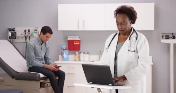 非洲中年医生在笔记本电脑上打字 年轻男性病人使用智能手机的诊所全景 高级黑人医生在手提电脑上记录单层拉丁裔病人 — 图库视频影像