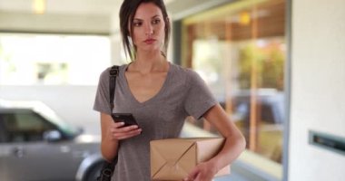 Evinin önünde duran bir kadın posta paketini tutarken cep telefonu kullanıyor. Milenyum kadını postaneye gitmeden önce akıllı telefon uygulamasını paketin izini sürmek için kullanıyor. 4k