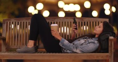 Kot ceketli hippi kız şehir caddesinde halka açık bankta yatıyor ve Bokeh Lights mesaj atıyor. Milenyum kadını kafasının altında sırt çantasıyla bankta uzanırken kahve tutuyor. 4k