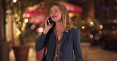 Orta yaşlı, beyaz bir kadın gece şehir merkezinde cep telefonu görüşmesi yapıyor. Gülen, gülümseyen profesyonel bir kadın akşam kaldırımda telefon görüşmesi yapıyor. 4k