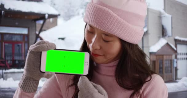 中国人女性の笑顔冬服を雪のダウンタウンで緑の画面でスマートフォンを保持 手袋とセーターで幸せな千年紀は 画面を表示し それを見て携帯電話を保持しています — ストック動画