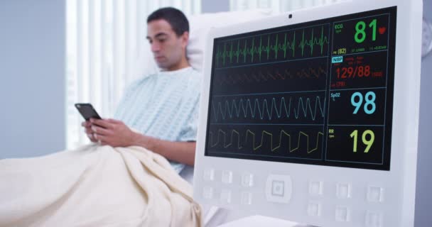 紧贴Ekg监视器 男病人躺在病床上 电话发短信 在病人通过智能手机阅读短信的同时 密切关注病人在监视器屏幕上阅读的基本信息 — 图库视频影像