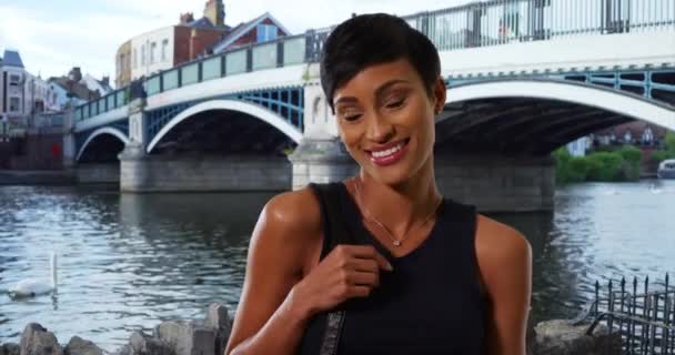 イギリスのウィンザーで財布を持つかなりアフリカ系アメリカ人女性の笑顔の肖像画 ウィンザータウンブリッジによってポーズ現代的なヘアカットを持つ魅力的な黒人女性を自信を持って — ストック動画