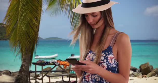 日当たりの良いビーチの設定で携帯電話上のルーマーとフェドーラのテキストメッセージの千年紀の女の子 メッセージを送信するために携帯電話を使用して熱帯休暇に彼女の20代の魅力的な女の子 — ストック動画