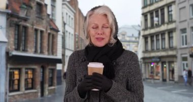 Soğuk sonbahar gününde Bruges 'de elinde sıcak bir fincan kahveyle mutlu bir turist. Kış kıyafetleri içinde olgun bir emeklinin Belçika 'da tatilde çay içmesi. 4k