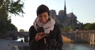 Paris, Fransa 'daki güzel siyahi kadın erkek arkadaşından gelen mesajları okuyor. Kahvesiyle birlikte şapkalı ve atkılı çekici genç bir kadın, Seine nehrinin kenarında akıllı telefon kullanıyor. 4k