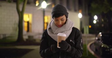 Soğuk sonbahar gecesinde mesaj atmak için eldivenini çıkaran 20 'li yaşlardaki sevimli siyahi kadın. Cep telefonuyla mesajlaşan, gülümseyen ve gülen Afro-Amerikan kadın portresi. 4k