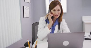 Güzel, orta yaşlı, beyaz bir doktorun cep telefonuyla konuşan ve ofisinde dizüstü bilgisayar kullanan portresi. Tıbbi hekimin hastasıyla telefonda test sonuçlarını tartışmasını kapatın.