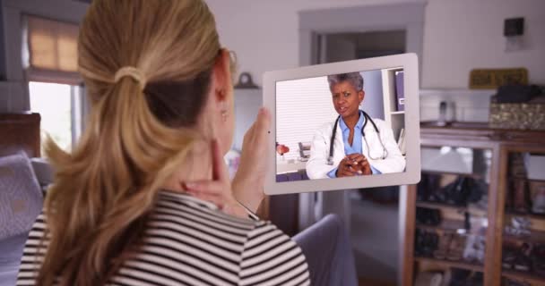 ベッドルームのタブレットで黒人女性医師と呼ばれる白人女性のビデオ 家庭からのビデオチャットを通じて医療専門家と話す後ろから示された女性または患者 — ストック動画