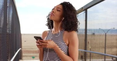 Çizgili bluzlu şirin zenci kadın portresi sahilde cep telefonuyla müzik dinliyor. Güzel Afrikalı kadın sıcak yaz gününde güzel bir şarkıyla dans ediyor. 4k