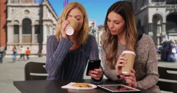 意大利威尼斯圣马可广场 一对快乐的女孩一边喝酒一边看手机 可爱的年轻的高加索女人看着圣马克大教堂前面的智能手机 — 图库视频影像