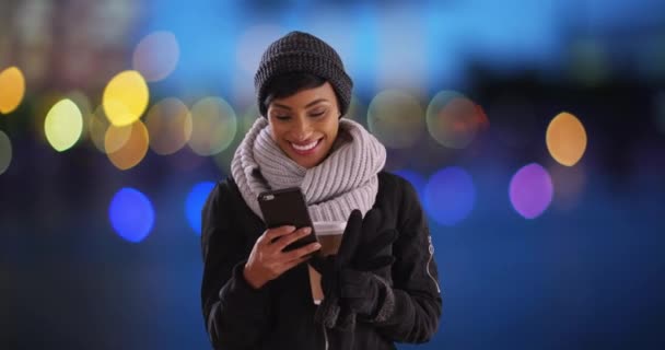 在户外用蓝色防盗灯在手机上用围巾和外套聊天的非洲裔美国女人很可爱 一个面带微笑的黑人女性的画像 在智能手机上看着什么 — 图库视频影像