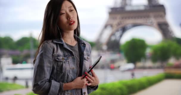 パリのエッフェル塔の前でスマートフォンやイヤフォンを片手に音楽を聴く若いヒップ ミレニアル ウーマン フランスの音楽に合わせて踊る素敵なアジアの女性のクローズアップ — ストック動画