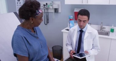 Yakışıklı genç Latin doktor hastane kliniğinde taşınabilir tabletle hasta notları alıyor. Orta yaşlı siyahi kadın, tıbbi geçmişi görmek için yüksek teknoloji not defteri kullanıyor.