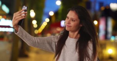 Şanzelize 'de gri kazaklı bir kadın cep telefonuyla selfie çekiyor. Dişi sırt çantalı, gece Paris caddesinde seyahat fotoğrafı çekiyor. 4k