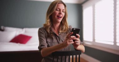 Beyaz kadın yatak odasında sandalyede oturan akıllı telefondan resimlere bakıyor. Evdeki cep telefonundan fotoğraflara bakan mutlu bir kadın. 4k