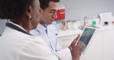 Elektronik bloknottaki röntgen ve boyun kemiklerine bakan genç Latin hasta ve kıdemli siyahi doktorların yakın çekimleri. Orta yaşlı Afrikalı doktorun portresi genç erkek hastaya omurga röntgeni sonuçları gösteriyor.