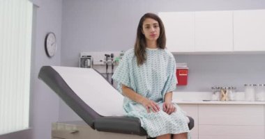Hastane kliniğinde yaslanmış tıbbi sandalyeye oturan sevimli Latin kadını yakından çek. Çekici, melez bir hastanın portresi. Klinikten kameraya bakıyor. Kasvetli bir ifade takınıyor.