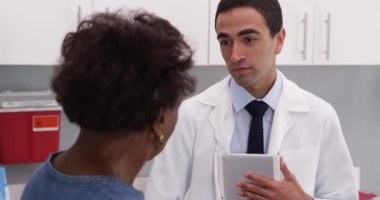 Hasta tıbbi geçmişini tartışmak için portatif tablet bilgisayar kullanan genç Latin doktorları kapatın. Çekici genç tıp doktoru yaşlı Afrikalı kadınla sağlık sorunları hakkında konuşuyor.