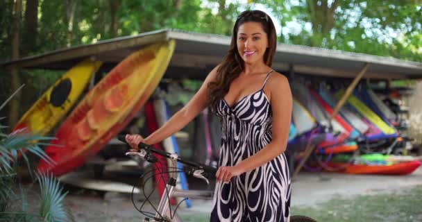 背景にサーフボードのレンタルと自転車で屋外に立ってファッショナブルなサンドレスで陽気なラティーナの女性 夏の日に外の自転車でポーズ休暇中の笑顔の女性の肖像画 — ストック動画