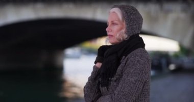 Paris tatilinde Seine nehrine bakan süveter ve eşarplı yaşlı bir kadın. Kış boyunca Fransa 'da seyahat eden yaşlı bir turist. 4k
