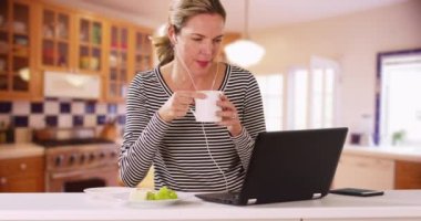 Beyaz kadın mutfakta oturuyor dizüstü bilgisayarla çalışıyor, müzik dinliyor, sabahları kahve içiyor. Evde bilgisayarda yazı yazan ve kahvaltı yapan bir kadın. 4k