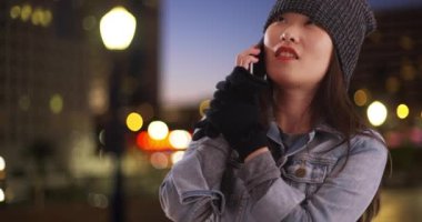 Cep telefonuyla konuşan ve Long Beach California 'da ısınmaya çalışan neşeli bir milenyum kadını. Mutlu genç Asyalı kadın gece şehir merkezinde akıllı telefondan sohbet ediyor. 4k