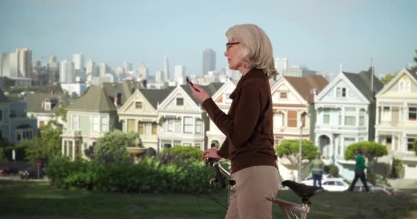 人気のアラモ広場の近所で電話で自転車のテキストメッセージで立って陽気な高齢者の女性のプロフィール サンフランシスコの有名な塗装女性の家の外のスマートフォン上の観光メッセージ — ストック動画