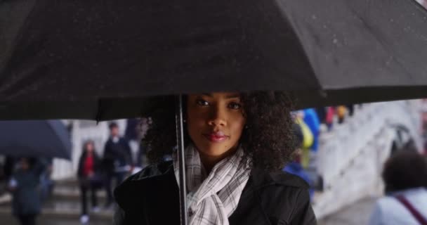威尼斯雨天站在雨伞下的非洲裔美国游客的近身 千禧年女人穿着黑色雨衣在街上等雨 同时游览意大利 — 图库视频影像